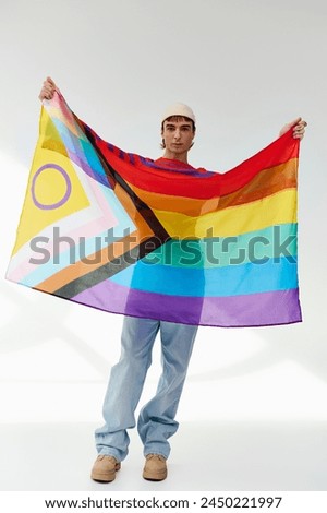 joyful appealing gay man in vivid outfit with white hat holding rainbow flag and looking at camera