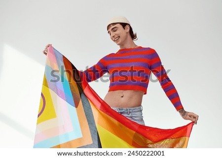 young appealing gay man in vibrant attire posing with rainbow flag and looking away on gray backdrop