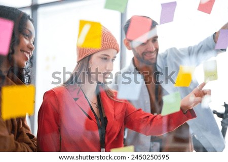 Business team uses a glass wall covered with sticky notes to outline processes during meeting. Engaged colleagues gather around a glass brainstorming board. plotting ideas and strategies. Royalty-Free Stock Photo #2450250595