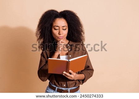 Portrait of intelligent minded woman with wavy hairstyle dressed brown shirt thoughtfully look at book isolated on beige color background