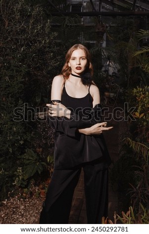 A female model stands illuminated against a background of plants. Dressed in a black jacket and top. Concept of fashion, stylish clothes and nature.
