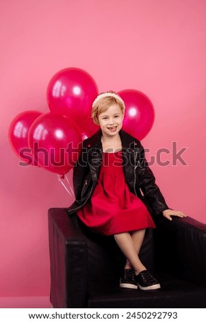 A child in a vibrant red dress and black leather jacket sits on a dark couch,surrounded by shiny pink balloons against a pink background.Concept for birthday parties, celebrations or fashion for kids.