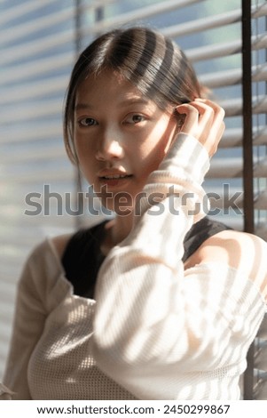A photo of a beautiful Asian woman with long dark hair wearing a white sweater, standing in front of a window with blinds.