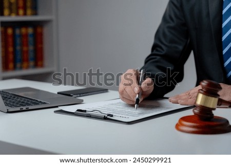 A lawyer in a suit is sitting at his desk, signing legal documents with a gavel on the side.