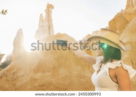 A woman in a cowboy hat is taking a picture of a mountain with her cell phone. The photo has a warm, sunny feel to it, and the woman's smile suggests that she is enjoying the moment