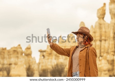A woman in a brown coat and hat is taking a picture of a mountain range. The photo has a sense of adventure and exploration, as the woman is capturing the beauty of the landscape