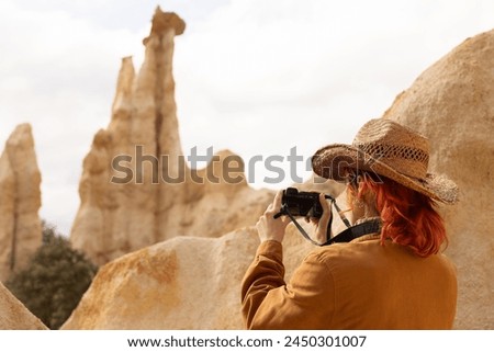 A woman wearing a cowboy hat is taking a picture of a mountain. The photo has a rustic, adventurous feel to it