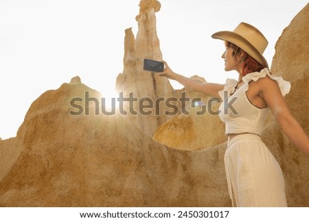 A woman is taking a picture of a mountain with a cell phone. The photo has a warm, sunny feel to it, and the woman is wearing a cowboy hat