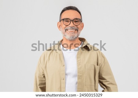 A cheerful senior man with a beard and glasses stands posing in a beige shirt against a gray background, exuding confidence and happiness