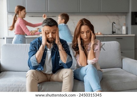 A stressed couple sits on a couch, looking exhausted, while their two children fight in the background of a modern home Royalty-Free Stock Photo #2450315345
