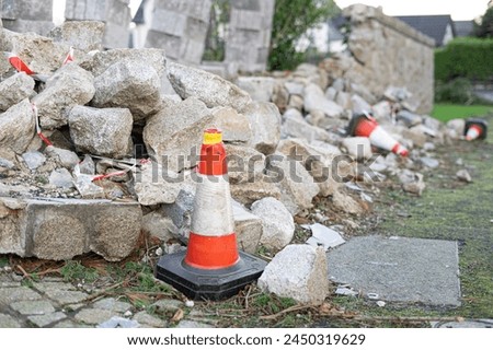 Stone wall fell over destroyed by storm tree being rebuilt rubble fallen on sidewalk front yard cobblestones warning orange white plastic traffic cone residential street road tape ground construction