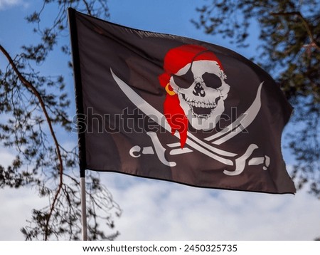 Black flag with white skull and red bandana flutters in the wind.  Pirate flag with Jolly Roger against the background of the spring blue sky in the forest. 