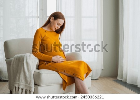 Pregnancy motherhood people expectation future. Pregnant woman with big belly sitting on chair near window at home. Girl hugging her tummy enjoying pregnancy. Maternity tenderness parenthood new life Royalty-Free Stock Photo #2450336221