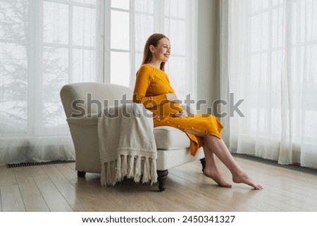 Pregnancy motherhood people expectation future. Pregnant woman with big belly sitting on chair near window at home. Girl hugging her tummy enjoying pregnancy. Maternity tenderness parenthood new life Royalty-Free Stock Photo #2450341327