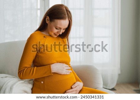 Pregnancy motherhood people expectation future. Pregnant woman with big belly sitting on chair near window at home. Girl hugging her tummy enjoying pregnancy. Maternity tenderness parenthood new life Royalty-Free Stock Photo #2450341331