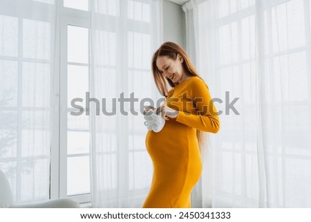 Pregnancy motherhood people expectation future. Pregnant woman with big belly holding newborn baby booties smiling at home. Young mom enjoying pregnancy. Maternity tenderness parenthood new life Royalty-Free Stock Photo #2450341333
