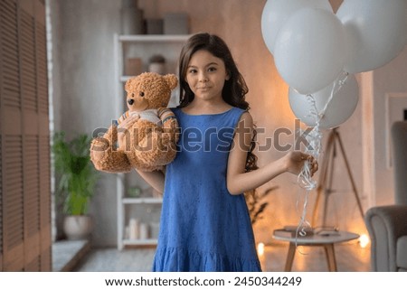 Happy cute little girl with long wavy brown hair standing posing with teddy bear toy gift present and white air balloons in her hand in blue pastel dress, cozy kid bedroom on background