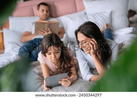 Cute little girl showing looking watching something to her mother in the tablet while lying in bed with father that reading book on the background. Family spending time weekend together at home