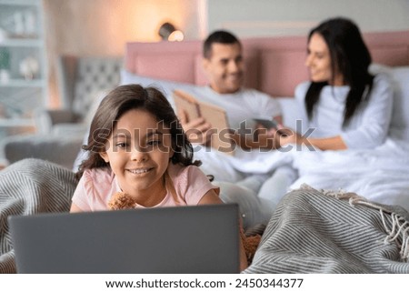 Smiling cute preteen little girl watching in notebook laptop while her father showing to her mother something in book on background in bed. Focus on daughter. Family lying in bed using gadget