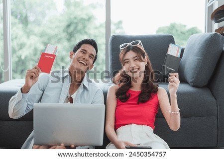 A couple is sitting on the floor with a laptop and tablet in front of them. They are smiling and seem to be enjoying their time together, holding passport and reservation hotel and ticket