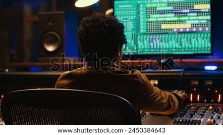 African american music producer mixing and mastering tracks on audio console, twisting knobs and pushing sliders to edit tunes. Sound engineer producing new songs in control room. Camera A.
