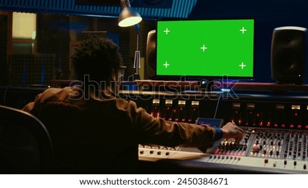 African american audio engineer uses greenscreen on pc in professional recording studio, working with mixing console and twisting knobs to edit tracks. Music producer creating tunes. Camera B.