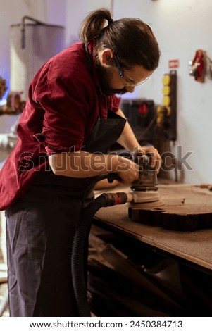 Craftsperson at work using orbital sander with fine sandpaper on lumber to achieve smooth finish. Woodworking expert in carpentry shop uses angle grinder on wood for professional results Royalty-Free Stock Photo #2450384713