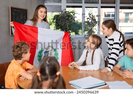 Excited young female teacher showing flag of Peru to schoolchildren preteens during history lesson in classroom Royalty-Free Stock Photo #2450396639