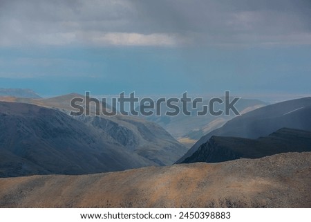Dramatic layered aerial view above large sharp ridge to sunlit green alpine deep valley among big cliffs far away in rain under gray cloudy sky. Mountain silhouettes on horizon in rainy low clouds.