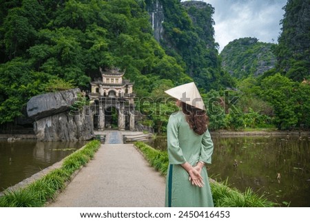 Back view of girl wearing Ao Dai in front of Outdoor park landscape with lake and stone bridge. Gate entrance to ancient Bich Dong pagoda complex. Ninh Binh, Vietnam travel destination