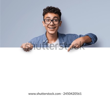 Excited happy young man wear glasses, braces stand behind, peep empty banner mockup sign board signboard, advertise show point sale slogan text place, isolated grey gray background. Dental care ad