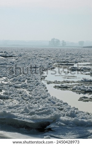 Thick ice blankets the River Elbe in Northern Germany, transforming its surface into a wintry wonderland. The frozen expanse reflects the pale sunlight, creating a serene and mesmerizing sight. Royalty-Free Stock Photo #2450428973
