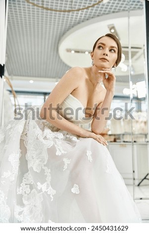 A young brunette bride in a flowing white dress sits elegantly on a chair in a wedding salon.