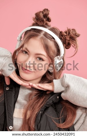 Pretty teenage girl with cute hair bumps on her head and wavy hair, listens to music on headphones. Pink background. Youth style.