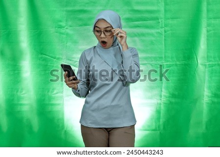 Shocked young Asian woman wearing hijab, glasses and blouse using mobile phone and taking of glasses isolated over green background