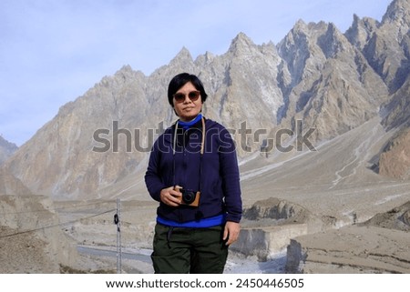 An Asian female tourist posing for a picture with the view of tall mountains during the fall season in Passu Valley, Gilgit-Baltistan, Pakistan.