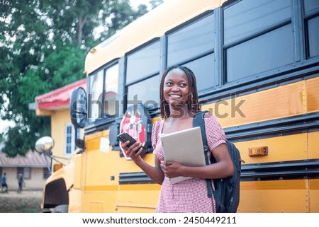 young Black female student dressed in a school uniform, standing confidently beside a yellow school bus. She is skillfully handling both a smartphone and a laptop, showcasing her ability to multitask Royalty-Free Stock Photo #2450449211