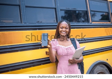 young Black female student dressed in a school uniform, standing confidently beside a yellow school bus. She is skillfully handling both a smartphone and a laptop, showcasing her ability to multitask Royalty-Free Stock Photo #2450449215