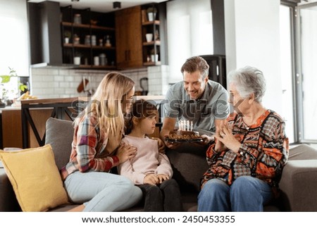 Heartwarming scene unfolds as a multi-generational family gathers on a couch to present a birthday cake to a delighted grandmother, creating memories to cherish Royalty-Free Stock Photo #2450453355