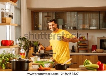 Indian young handsome man in kitchen taking selfie picture while cooking, using tablet pc and having coffee