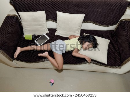 A relaxed young girl rests comfortably on a couch, engrossed in a digital tablet. A reusable water bottle sits beside her.