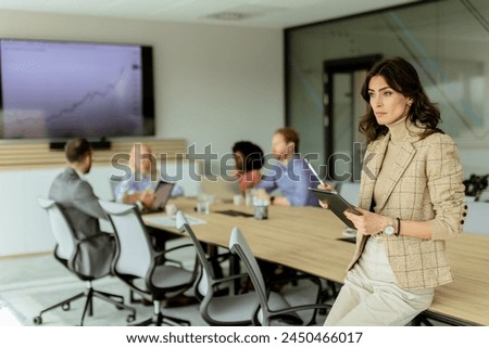 A poised woman with a tablet stands at the forefront of a collaborative office space, her team engaged in discussion behind her. Royalty-Free Stock Photo #2450466017