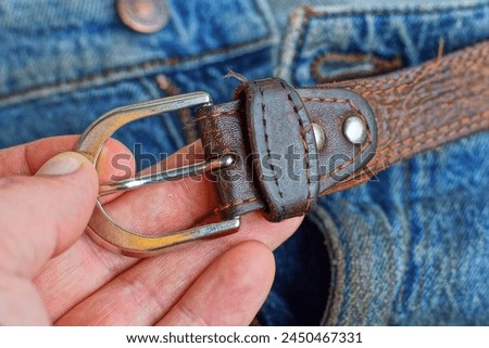 leather brown old belt with an iron buckle in hand on the background of blue stylish dirty fashionable men's jeans with an unzipped zipper