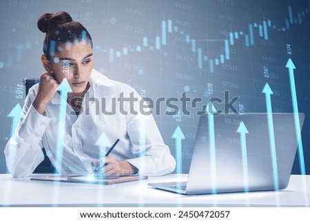 Attractive young businesswoman at desk with laptop and growing blue vertical arrows and candlestick forex chart on blurry index grid background. Economic growth and increase concept. Double exposure