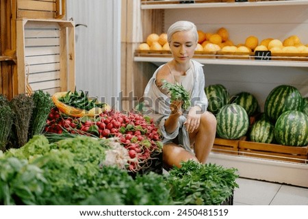 A young woman with short hair examining a bunch of greens while shopping for fresh produce in a well-stocked grocery store.