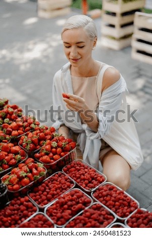 A woman in a summer outfit handpicks ripe strawberries from a vibrant selection of fresh berries at a local market. Royalty-Free Stock Photo #2450481523