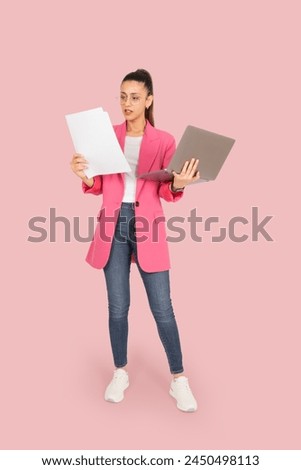 Focused businesswoman, full body length brunette caucasian focused businesswoman. Holding papers and laptop. Female executive analyzing paperwork using computer. Pink jacket, jeans and sneakers.