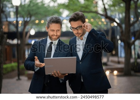 Two businessmen on the street, deeply engrossed, eyes fixed on their laptop screen, perhaps discussing a crucial deal or strategizing for their next big project in the bustling cityscape.
