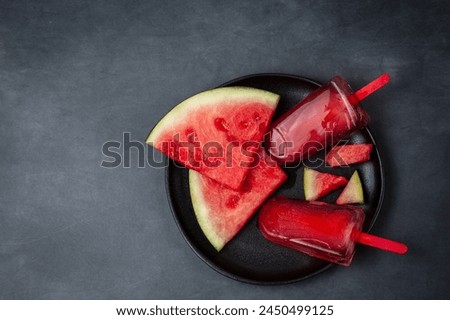 Aerial view of black plate with popsicles and pieces of watermelon on gray table, horizontal, with copy space