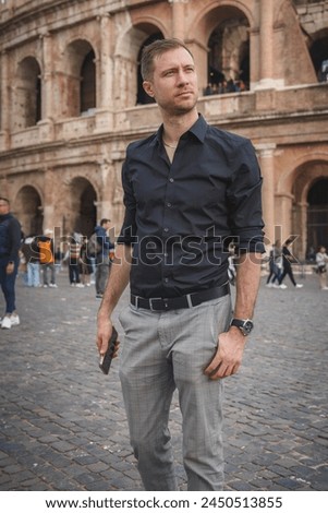 Fashionable man in smart attire holding sunglasses, standing in front of iconic Colosseum in Rome, Italy. Historical location with blurred background of tourists.